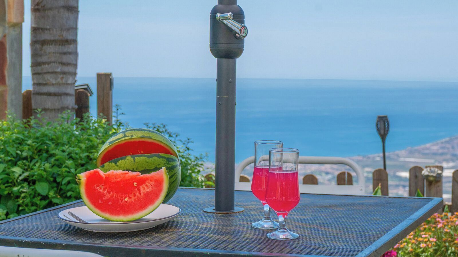 Ein Tisch mit einer Wassermelone, zwei Gläsern pinkfarbenen Getränks und einem Wasserhahn, mit Blick auf das Meer.