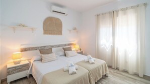 A double bed with two pillows, a bedside table with a lamp, a wall-mounted air conditioner, and a window with curtains.