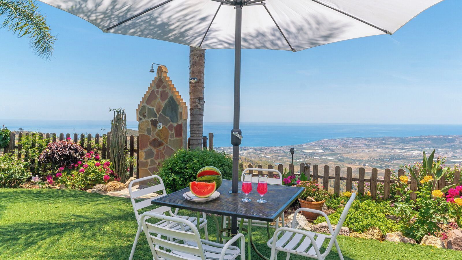 A patio with a table, chairs, a watermelon, and two glasses of red liquid, overlooking a scenic view.