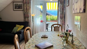 Ein gemütliches Wohnzimmer mit einem schwarzen Sofa, Holzstühlen, einem runden Tisch mit einer blumigen Tischdecke, einem Schachbrett und einer Vase mit Blumen.