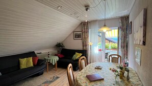 Ein gemütliches Wohnzimmer mit einer schwarzen Couch, einem kleinen Tisch mit grüner Tischdecke und einem Esstisch mit einer blumigen Tischdecke.