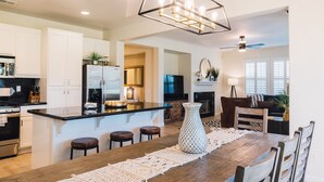 A modern kitchen with white cabinets, a central island with stools, a chandelier, and a dining area with a wooden table and chairs. - Golf Course Luxury with Pool, Spa, Casita & Mountain Views! (Indio)