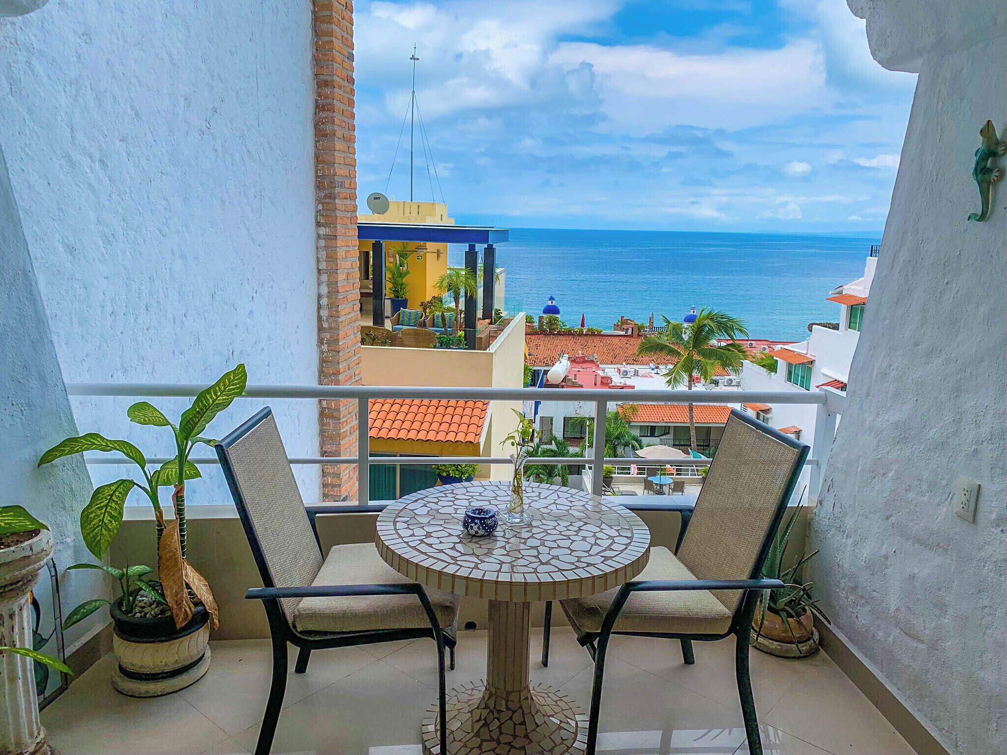 A balcony with a round table and two chairs, overlooking a coastal view with buildings and palm trees.
