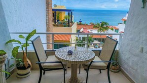 A balcony with a round table and two chairs, overlooking a coastal view with buildings and palm trees.