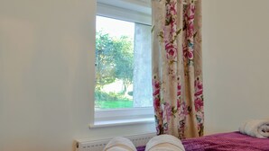 A bedroom with a bed, a window with floral curtains, and a view of greenery outside. - Cornel Clyd (Cosy Corner) (Felinfach, near Lampeter)