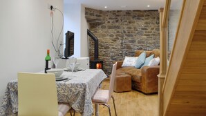 A cozy living room with a stone wall, a fireplace, a television, and a dining area with a table set for two. - Cornel Clyd (Cosy Corner) (Felinfach, near Lampeter)