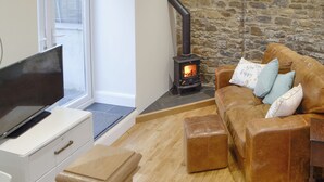 A cozy living room with a stone wall, a fireplace, a leather sofa with cushions, a television on a white cabinet, and a wooden floor. - Cornel Clyd (Cosy Corner) (Felinfach, near Lampeter)