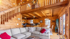 Interior acogedor de una cabaña de troncos con un amplio sofá seccional en forma de L, una escalera, una zona de cocina y una mesa de comedor.