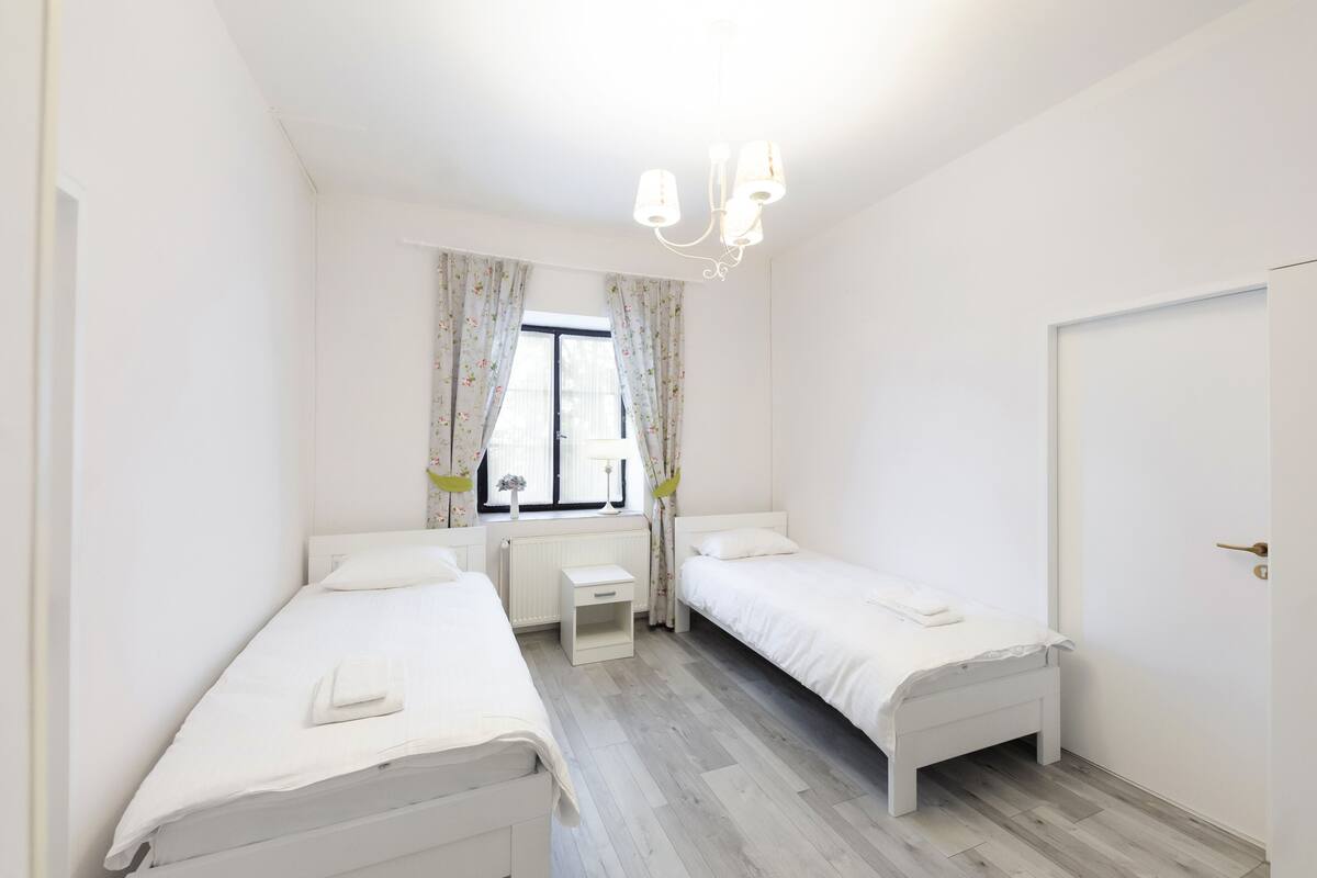 A bedroom with two single beds, a small desk, a window with curtains, and a chandelier.