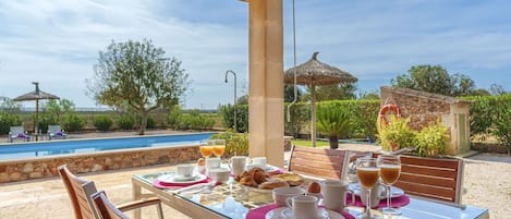 An outdoor dining area with a table set for breakfast, overlooking a swimming pool and garden.