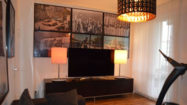 Uma sala de estar com televisão de tela plana, duas luminárias, uma esteira e uma parede decorada com várias molduras.