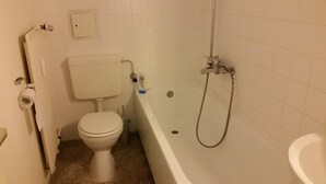Uma casa de banho com sanita, banheira com chuveiro e suporte para papel higiénico.