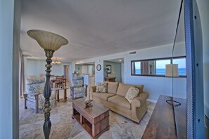 A spacious living room with a beige sofa, a wooden coffee table, and a decorative lamp. There is a dining area with a table and chairs, and a view of the ocean through large windows. - Seachase W 101 2 Bedroom 2 Bath unit. Beach Chair Service for 4 Included (Panama City Beach)