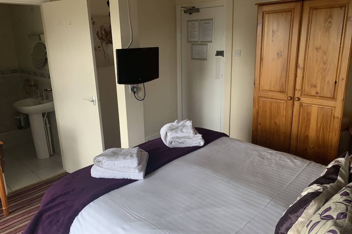 A hotel room with a bed, a wooden wardrobe, a television mounted on the wall, and a bathroom visible through an open door.