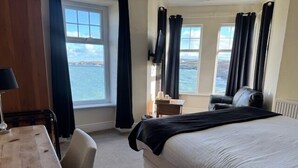 A hotel room with a bed, a desk, a chair, and a view of the sea through large windows. - The Trecastell Hotel (Amlwch)