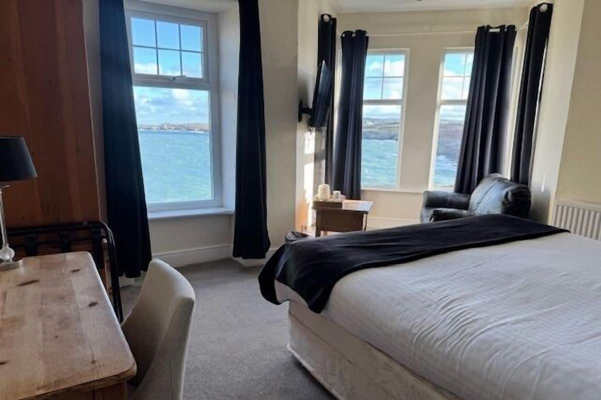 A hotel room with a bed, a desk, a chair, and a view of the sea through large windows.