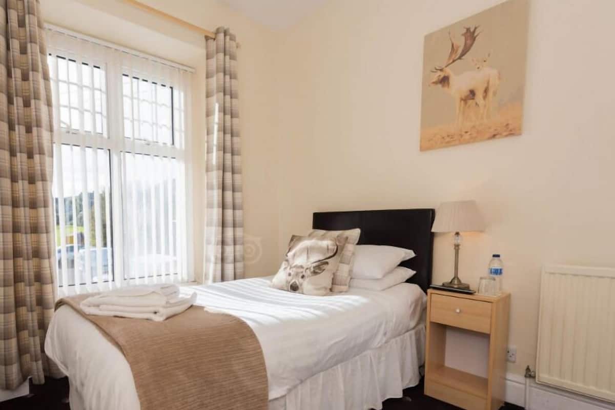 A neatly made bed with a white duvet and a beige throw blanket, a bedside table with a lamp and a bottle, and a painting of a deer on the wall.