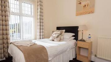 A neatly made bed with a white duvet and a beige throw blanket, a bedside table with a lamp and a bottle, and a painting of a deer on the wall.