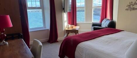 Habitación de hotel con cama, escritorio, silla, una mesita y vistas al mar a través de amplias ventanas.