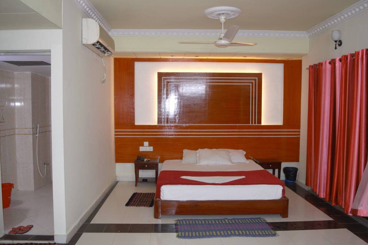 Habitación de hotel con cama, mesita de noche, ventilador de techo, ducha y ventana con cortinas.