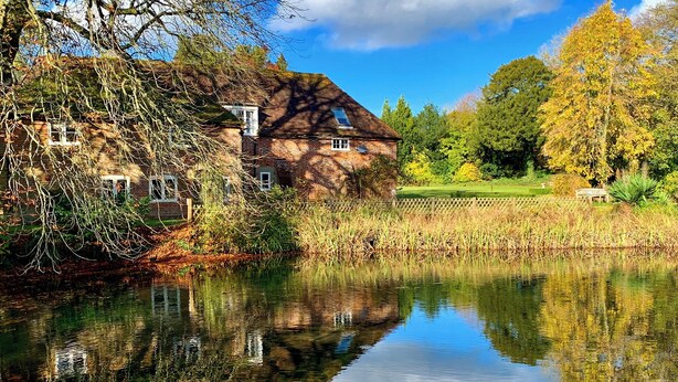Een rustiek huis aan een kalm meer, met een heldere spiegeling, omringd door bomen en een gazon.