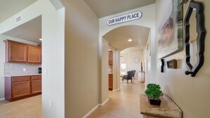 A spacious hallway with a sign reading "OUR HAPPY PLACE," leading to a kitchen area with wooden cabinets and a dining area with a chair and a potted plant. - *Tranquil Living in the City* (Las Cruces)