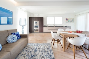A modern living room with a sofa, a dining table with chairs, a kitchen area with cabinets and appliances, and a large window providing natural light.