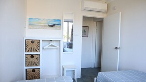 Une chambre avec deux lits, une armoire contenant des paniers en osier, une petite table et un tableau représentant un bateau accroché au mur.