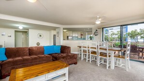 A modern living room with a brown sofa, blue cushions, a wooden coffee table, and a dining area with white chairs and a wooden table. - Peninsula Waters, 3/2-4 Soldiers Point Rd - Beautiful Air Conditioned Unit with Pool, Lift & WIFI (Soldiers Point)