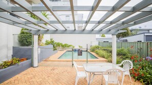 Área externa coberta com piscina, cadeiras brancas e uma mesa.