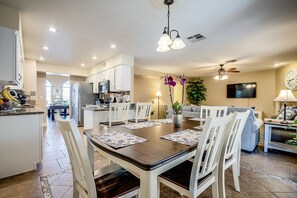 A modern kitchen with white cabinets, a dining area with a wooden table and white chairs, a ceiling fan, and a TV mounted on the wall. - Upscale 4 BR Home with RV parking (Henderson)