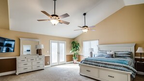 Une chambre à coucher comprenant un lit, deux ventilateurs de plafond, une commode, un téléviseur, une plante et une fenêtre avec des stores.
