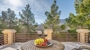 Une table ronde en osier, sur laquelle sont disposés un plateau de fruits et deux verres à vin, se trouve sur un balcon offrant une vue sur les arbres et les montagnes.