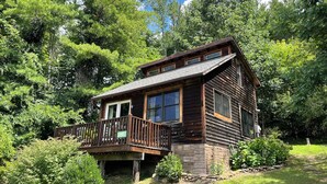 A rustic cabin with a wooden deck, surrounded by lush greenery and trees.