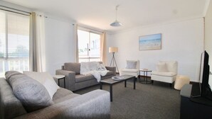 A modern living room with a grey sofa, a white armchair, a small coffee table, and a floor lamp. There is a large window with blinds, a painting on the wall, and a flat-screen TV on a stand.
