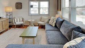 A living room with a grey sofa, a small round table, and a white armchair. There is a window with blinds and a decorative wall piece. - Mountains to Sea | Pet Friendly + Steps to Beach! (Holly Ridge)