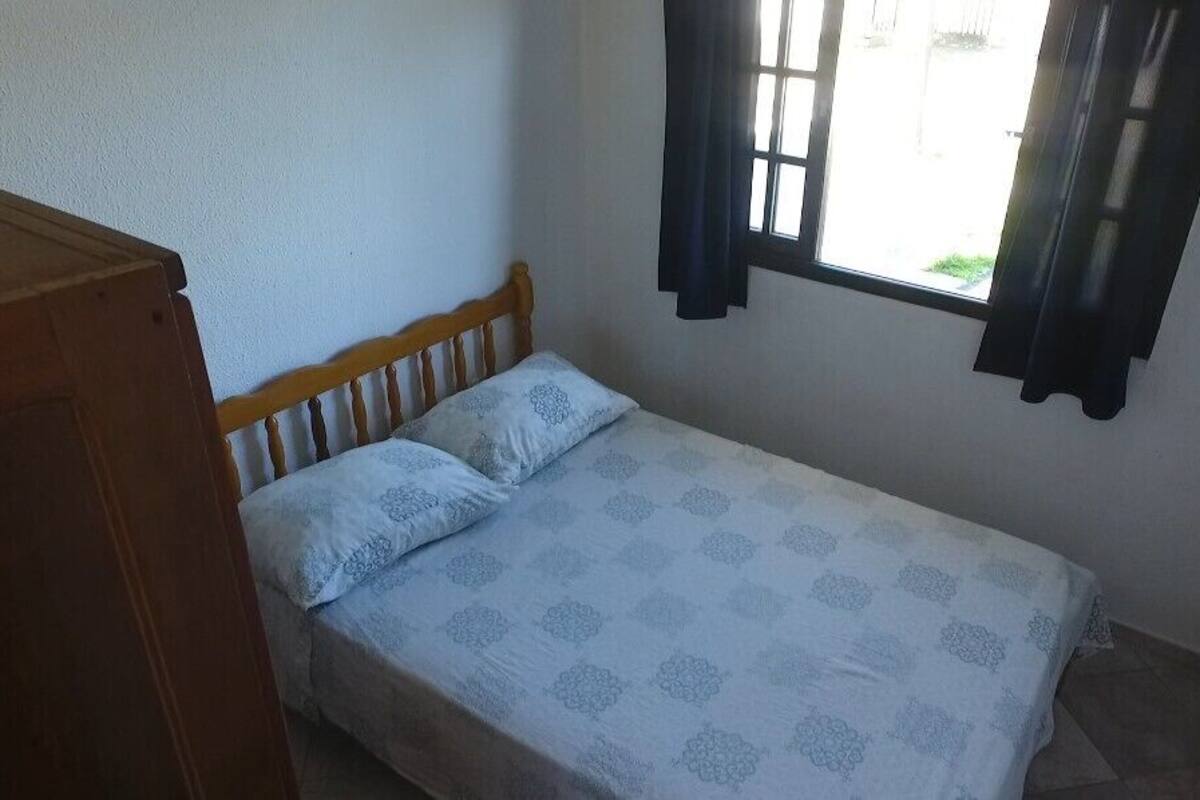 Ein kleines Zimmer mit einem Einzelbett, einem Holzkopfstück, einem Fenster mit Vorhängen und einer schlichten Wand.