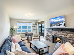 A living room with a stone fireplace, a large TV, and a view of the outdoors through a sliding glass door. - Contemporary Design w/Balcony-Seasons at Sandpoint (Sandpoint)