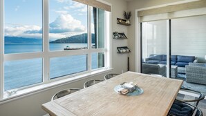 Un coin repas avec une grande fenêtre donnant sur un plan d’eau et des montagnes. On y trouve une table avec des chaises et un canapé à l’arrière-plan.