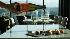 A hand pouring white wine into two glasses on a table with macarons, in a room with a view of mountains through large windows. - Craftsman on Whitman · Updated Craftsman on Whitman, Close to Everything! (Walla Walla)