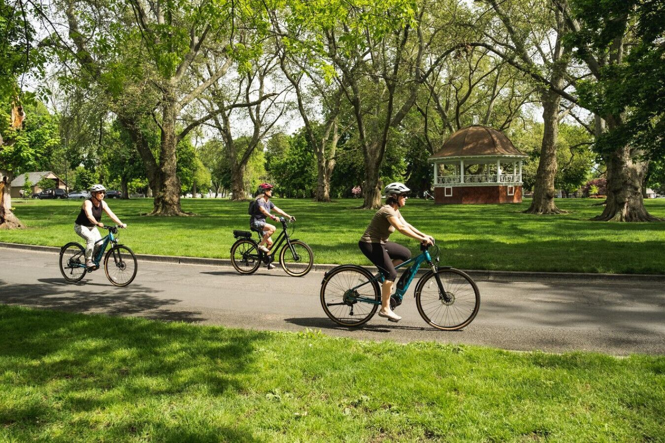 Three cyclists on a paved path in a park with trees and a gazebo in the background.