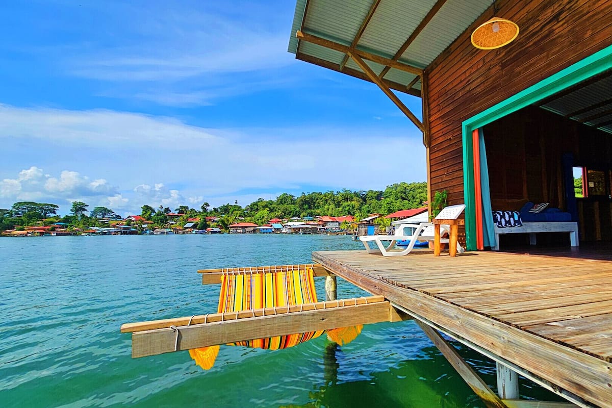 A wooden dock extending into clear blue water, leading to a building with a covered porch and outdoor seating.