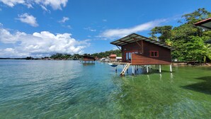 A wooden house on stilts over clear water, with a view of the surrounding landscape and other buildings.