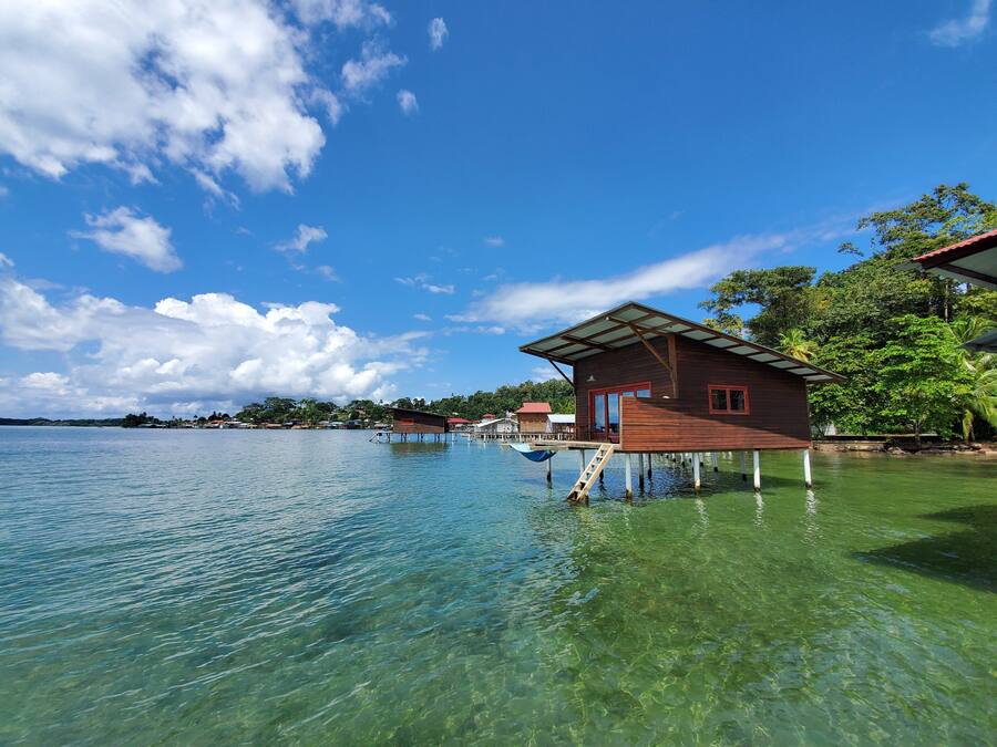 A wooden house on stilts over clear water, with a view of the surrounding landscape and other buildings.