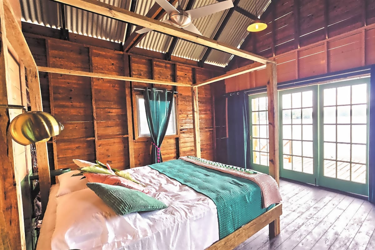 A rustic wooden room with a four-poster bed, a ceiling fan, and large windows.