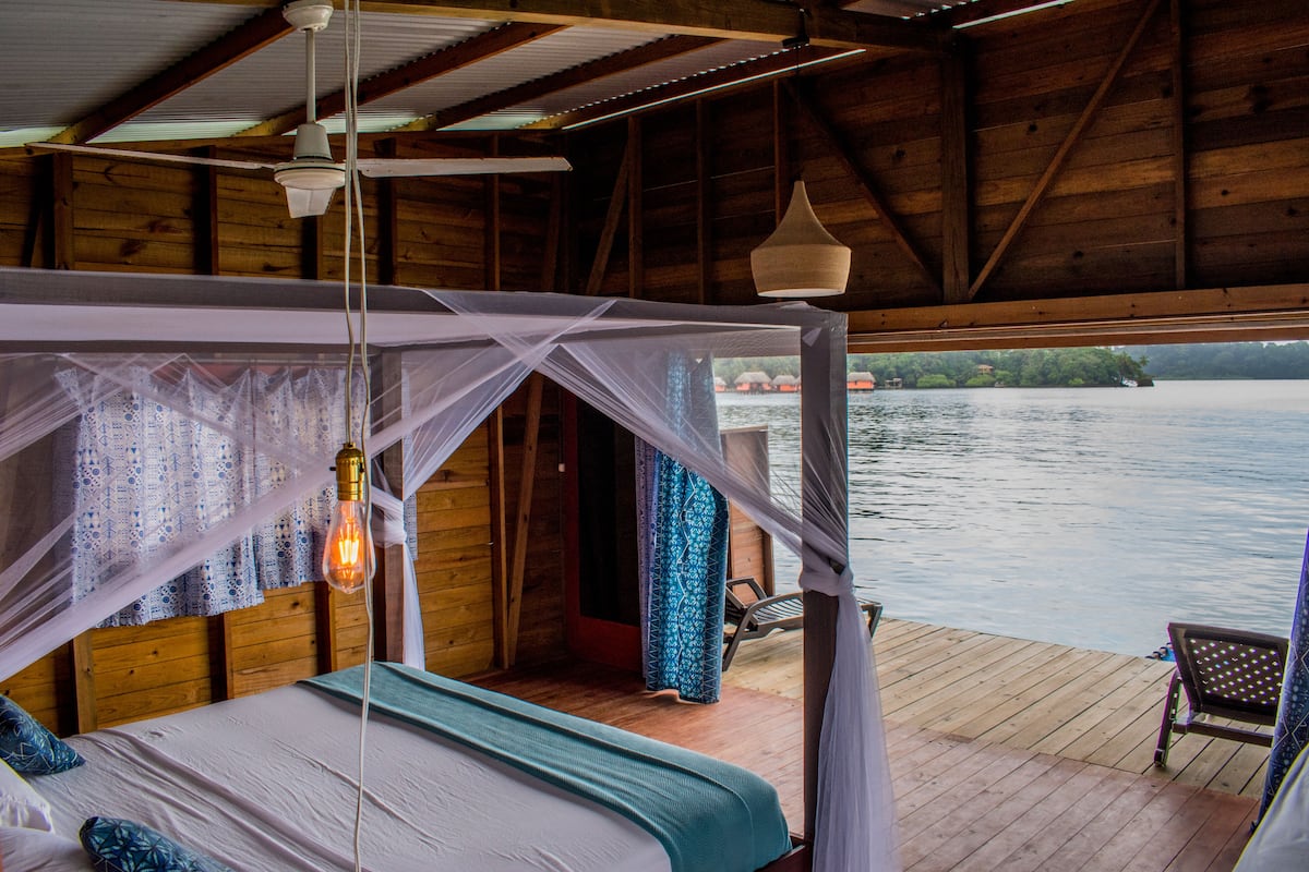 A wooden cabin with a bed under a mosquito net, a wooden deck, and a view of a lake.