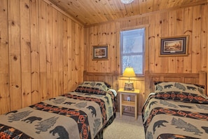 Quarto com paredes de madeira, duas camas com roupa de cama estampada, uma mesa de cabeceira com abajur e quadros nas paredes.