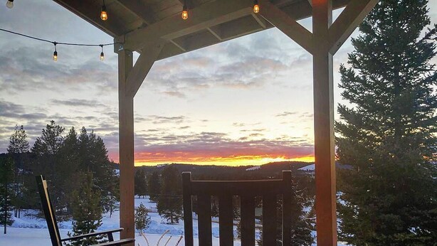 A wooden deck with two rocking chairs and a table, overlooking a snowy landscape with a sunset. - Georgetown Lake Retreat (Anaconda)