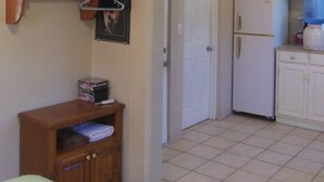 A compact room with a bed, a small wooden cabinet, a television mounted on the wall, a door, and a kitchenette area with a refrigerator and a water dispenser.