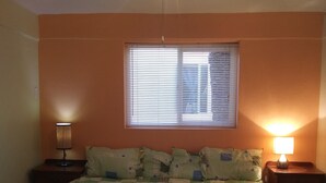 A bedroom with a bed, two bedside tables, a ceiling fan, a window with blinds, and a wall lamp.